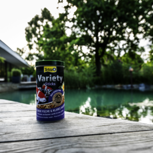 Bahçe havuzu kenarında ahşap zemin üzerinde duran Tetra Pond Variety Sticks balık yemi kutusu
